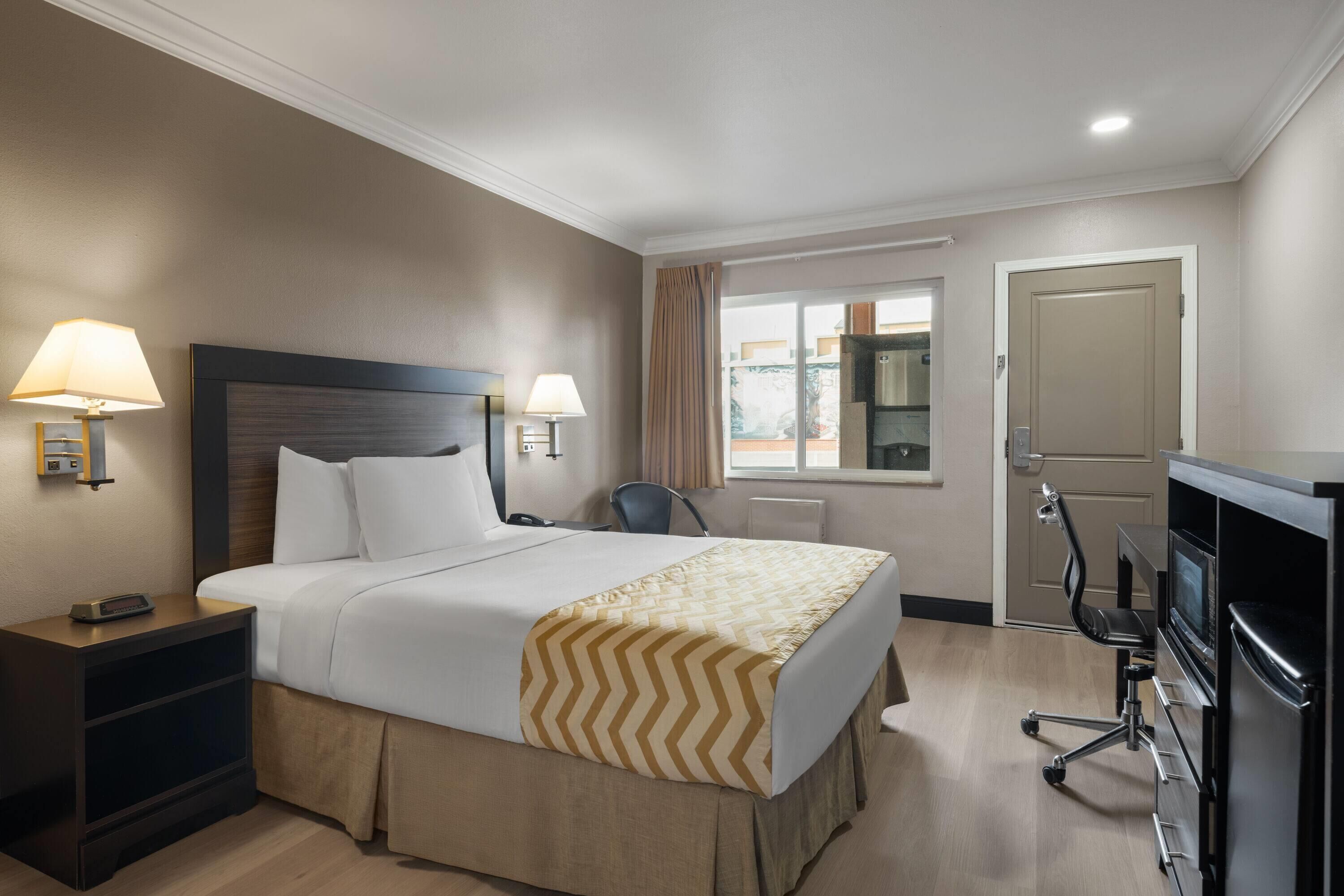 Una habitación de hotel con cama, lámparas de noche, un escritorio con ordenador, una silla y una puerta.