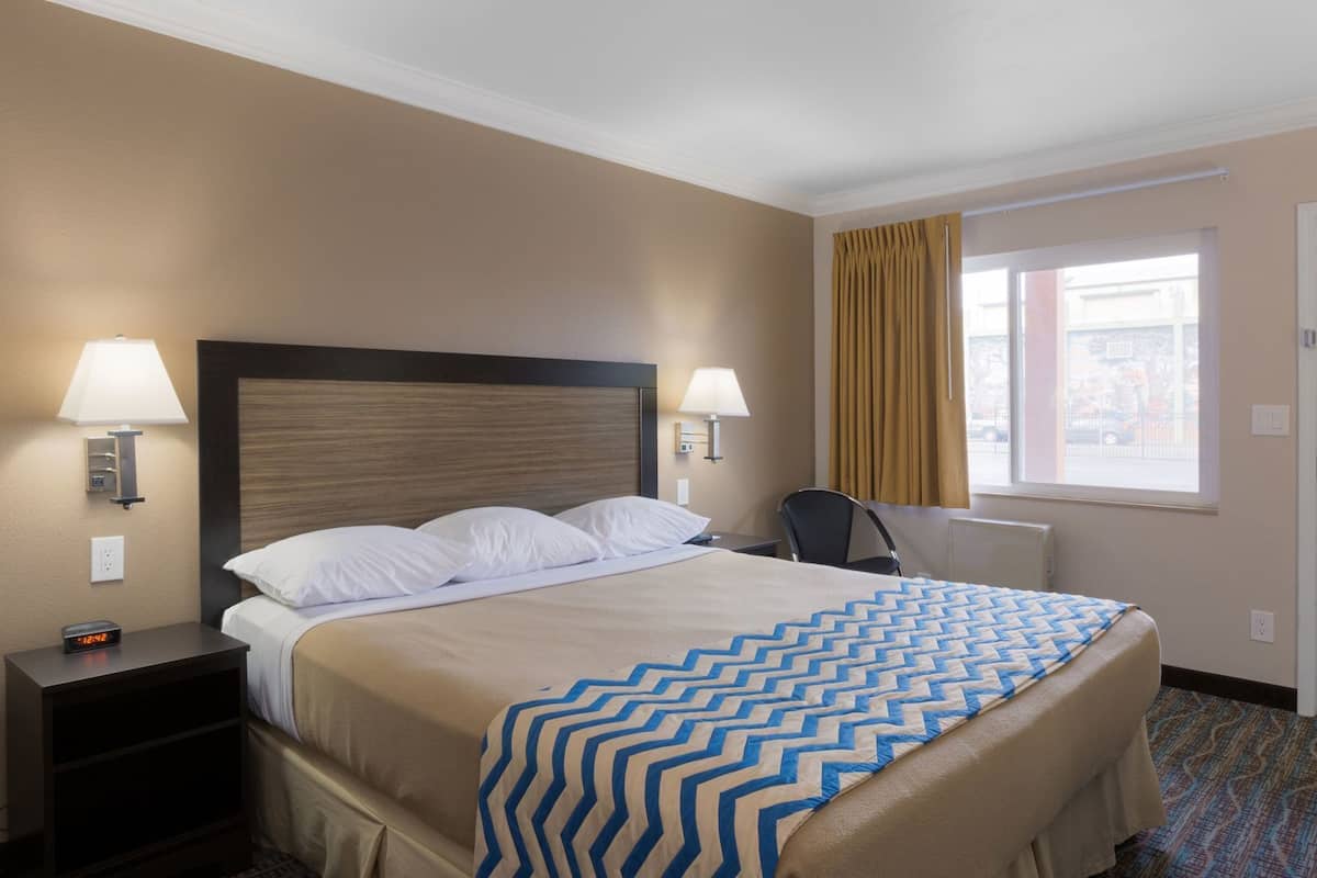 A hotel room with a bed, two bedside lamps, a chair, a window with curtains, and a nightstand with a clock.