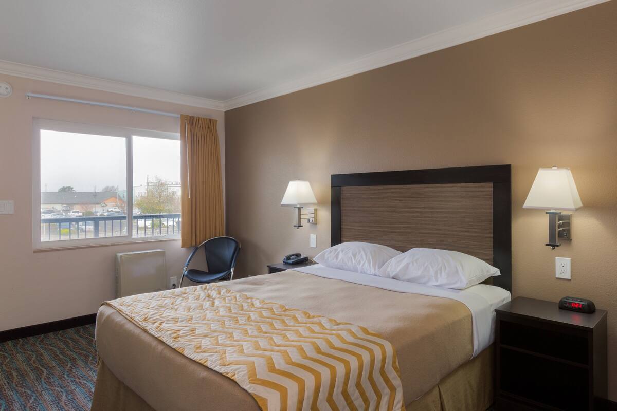 A hotel room with a large bed, two bedside lamps, a chair, a nightstand, and a window with a view of the outside.
