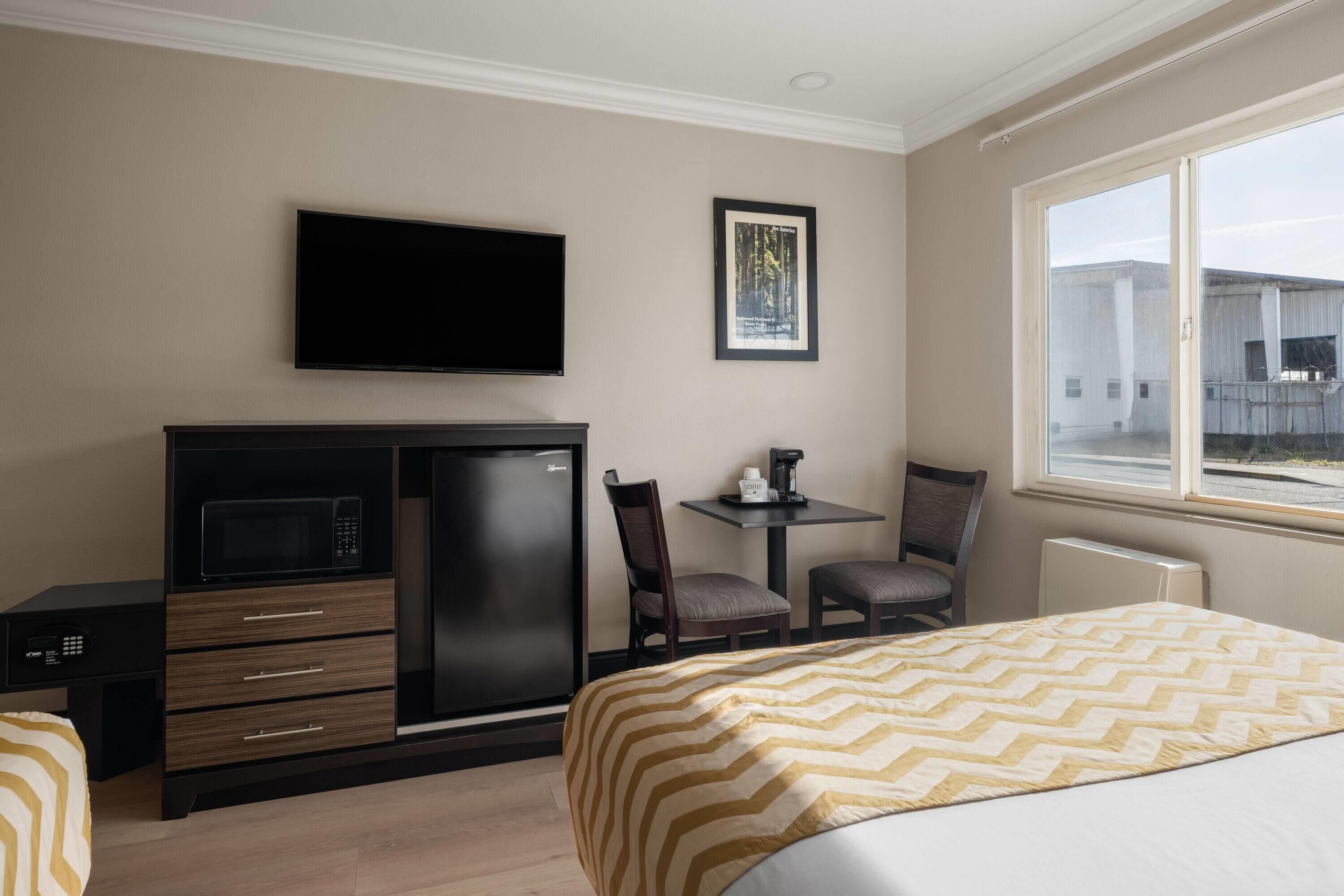 A hotel room with a bed, a desk with a microwave, a refrigerator, a TV, a framed picture, and a window.