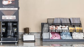 Une machine à café proposant différentes options : café normal, eau chaude et café décaféiné. À côté, on trouve un présentoir avec des articles de toilette et un panier rempli de magazines.