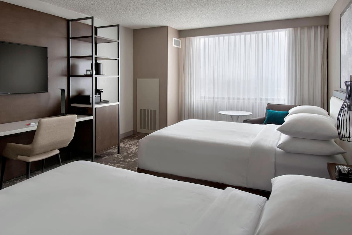 A hotel room with two beds, a desk with a chair, a TV, a small table, and a window with curtains.