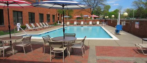 A poolside area with lounge chairs and umbrellas, a brick building in the background, and a clear sky.