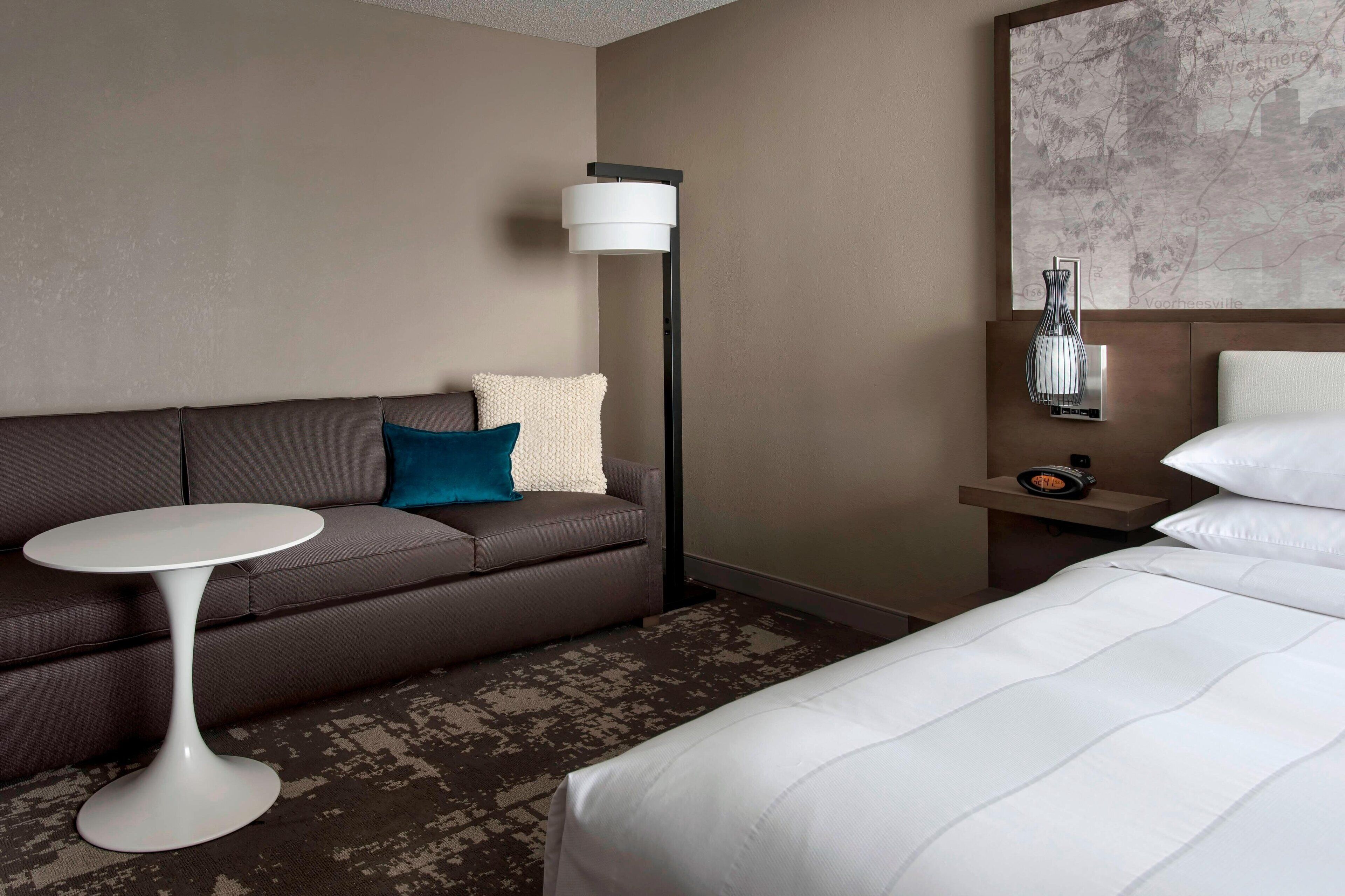 A hotel room with a bed, a sofa, a small round table, a floor lamp, and a nightstand with a clock.