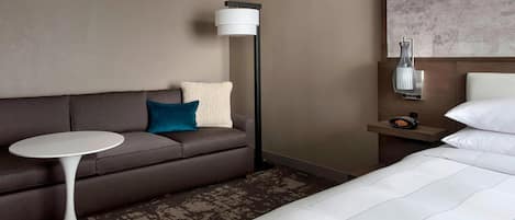 Habitación de hotel con cama, sofá, una mesita redonda, lámpara de pie y mesita de noche con un reloj.