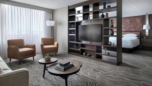 A hotel room with a large flat-screen TV, two armchairs, a sofa, a coffee table, a bookshelf, and a bed with a headboard. - Marriott Albany (Albany)