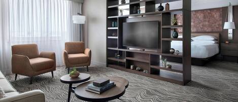 A hotel room with a large flat-screen TV, two armchairs, a sofa, a coffee table, a bookshelf, and a bed with a headboard.