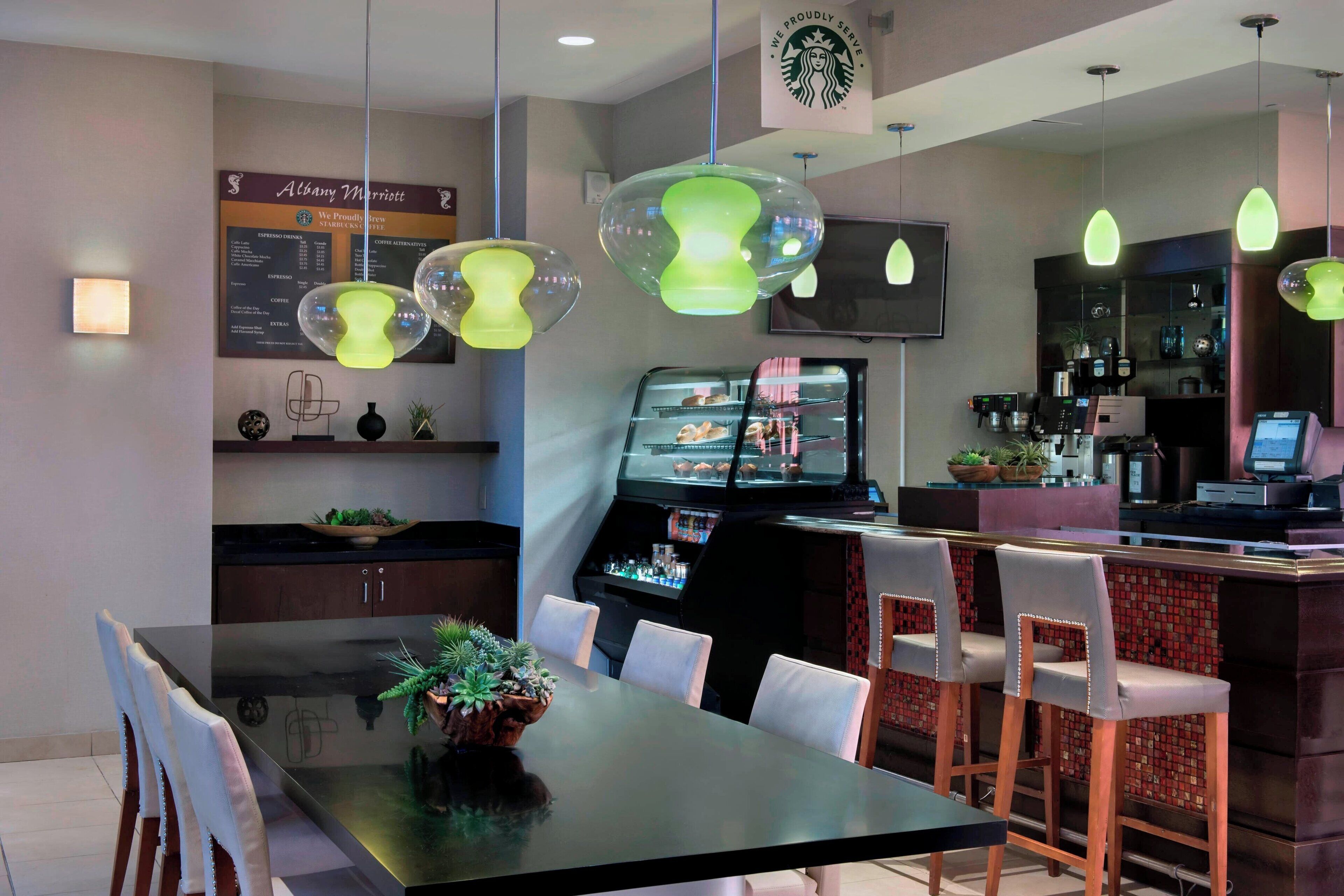 A modern hotel restaurant with a dining area featuring a large table and chairs, a bar area with stools, and a coffee machine.