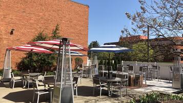 An outdoor patio area with tables and chairs, two large umbrellas, and a brick building in the background.