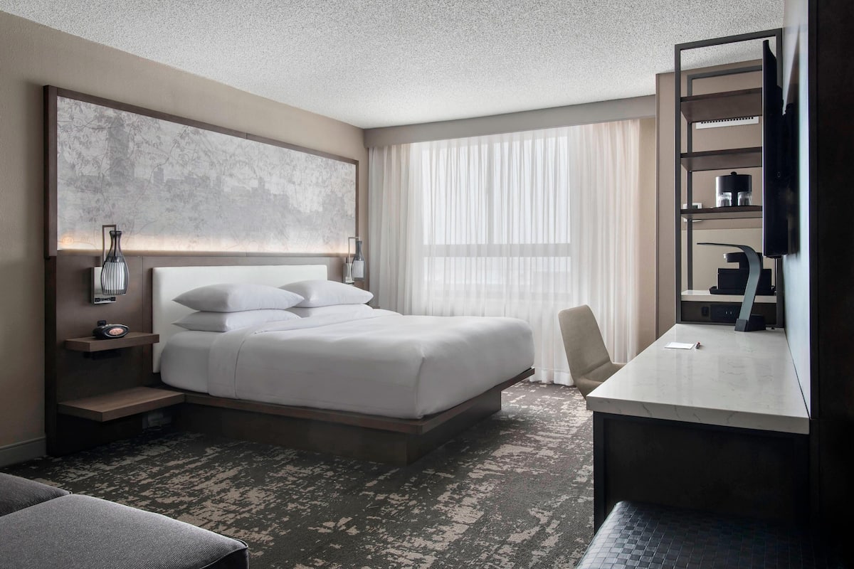 A modern hotel room with a large bed, a desk, a chair, a TV, and a bookshelf.