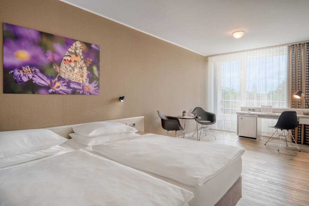 Habitación de hotel con dos camas, un escritorio con silla, una mesita y un ventanal grande con cortinas.