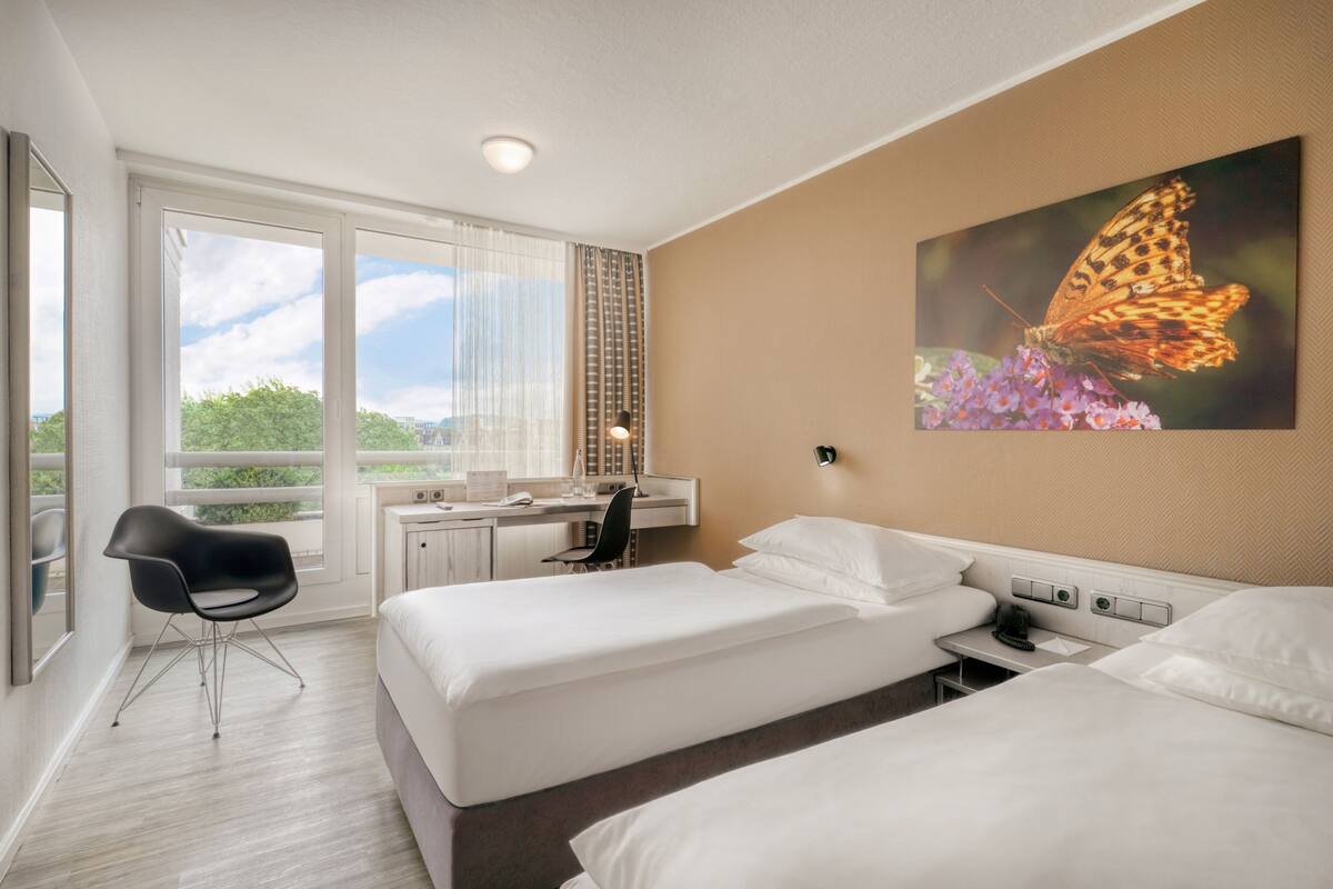 Habitación de hotel con dos camas, un escritorio, una silla y un amplio ventanal con vistas a zonas verdes.