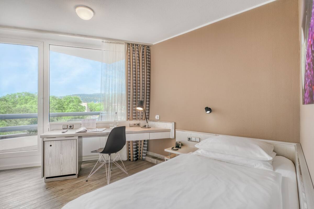 Habitación de hotel con cama, escritorio, silla y un amplio ventanal con vistas a zonas verdes.