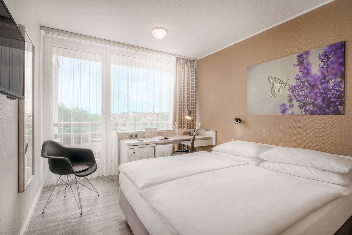 Habitación de hotel con cama, escritorio, silla y un amplio ventanal con cortinas.