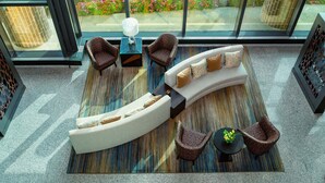 A modern lounge area with a curved sofa, two armchairs, a small table with a lamp, and a view of a flower garden through large windows. - The Westin Southfield Detroit (Southfield)