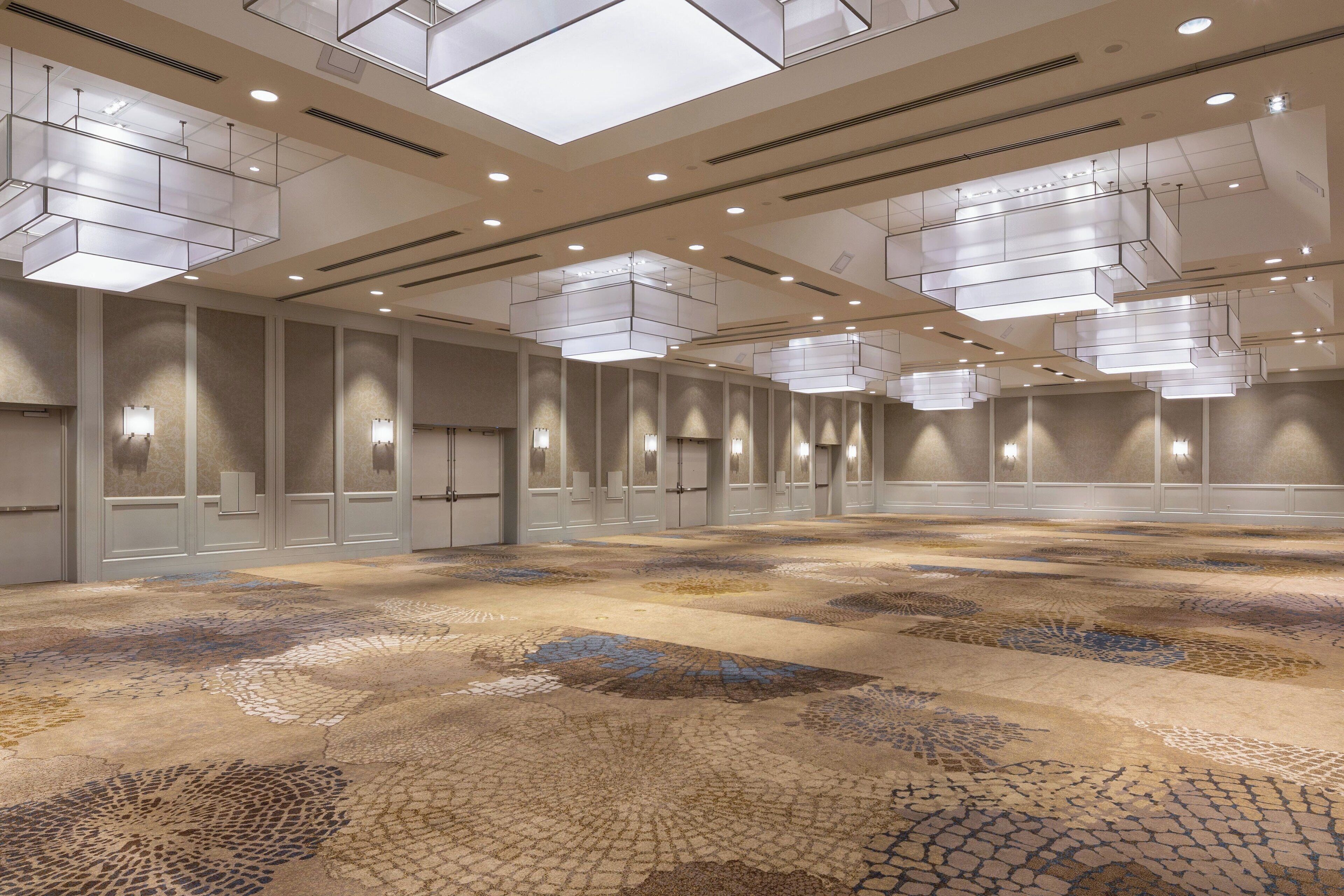A spacious hotel lobby with a patterned carpet, recessed lighting, and a high ceiling with hanging fixtures.