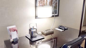 Habitación de hotel con escritorio de vidrio, cafetera, teléfono y un cuadro en la pared.