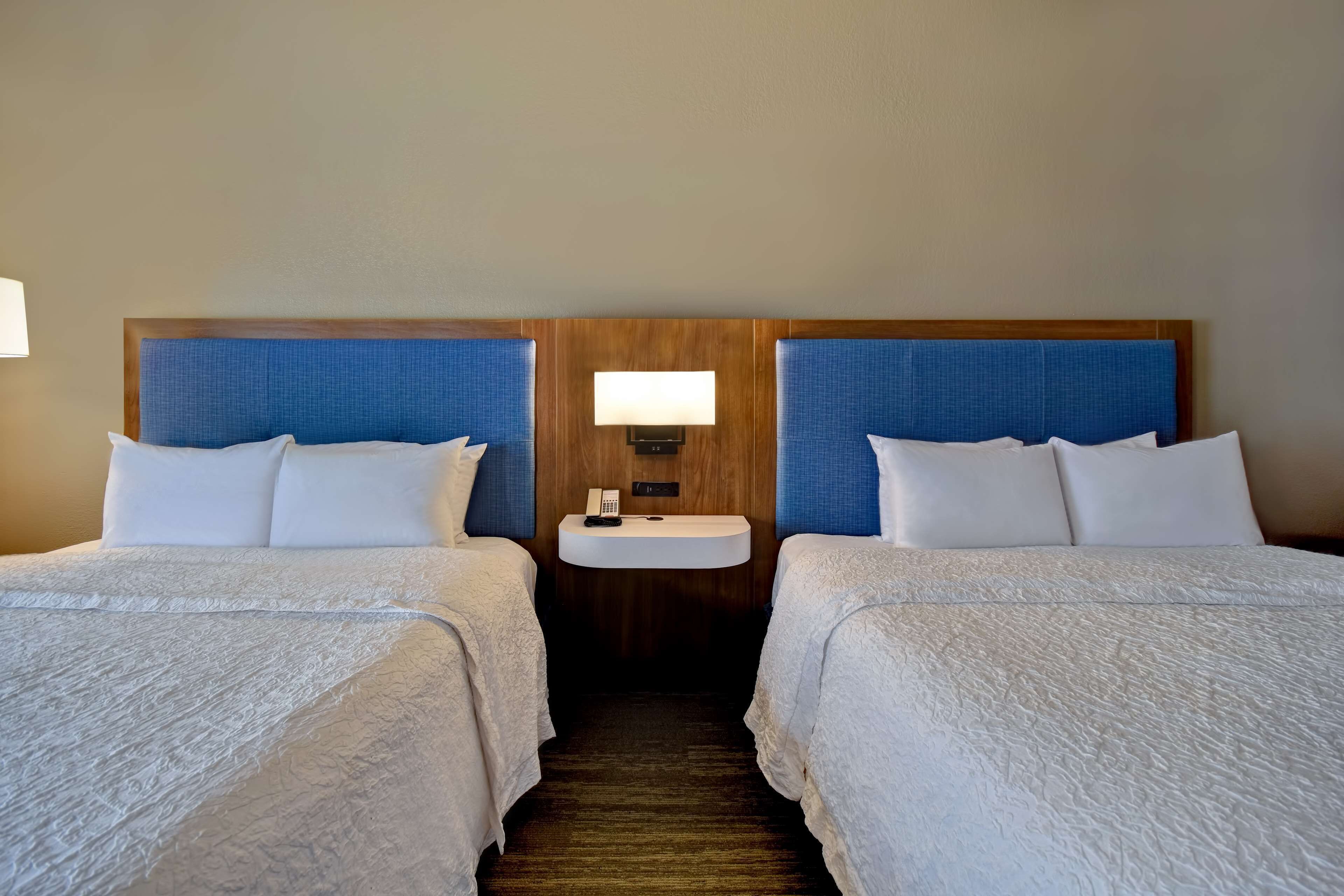 Una habitación de hotel con dos camas, una mesita de noche pequeña con una lámpara y un cabecero de madera.