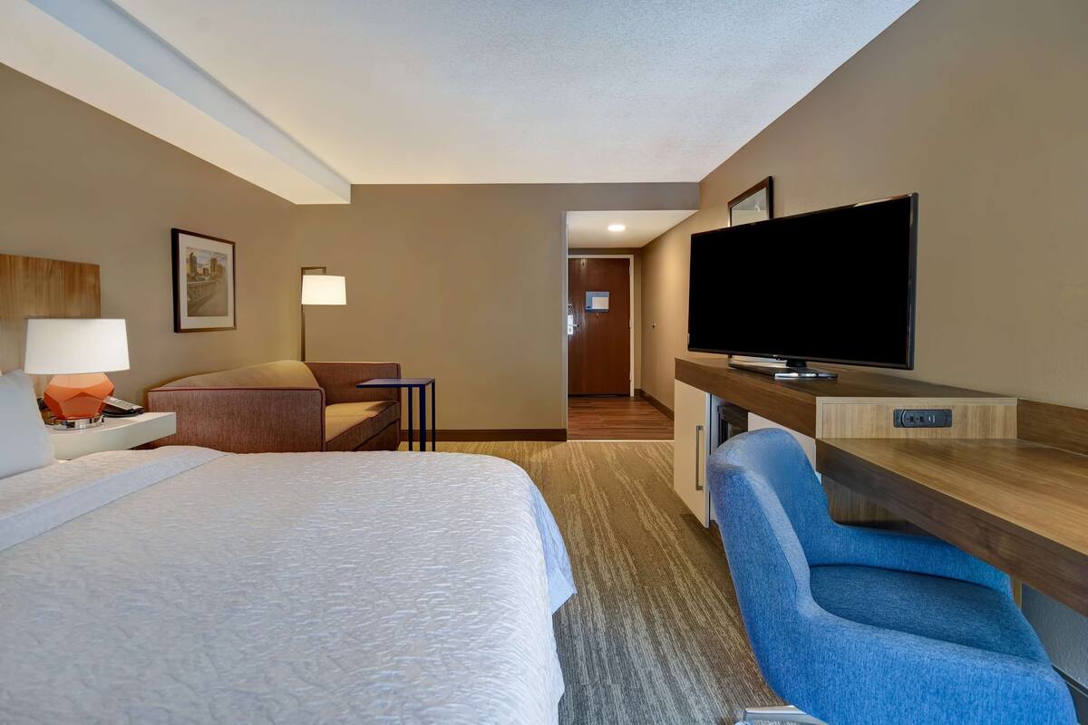 A hotel room with a large bed, a desk with a flat-screen TV, a chair, and a lamp.