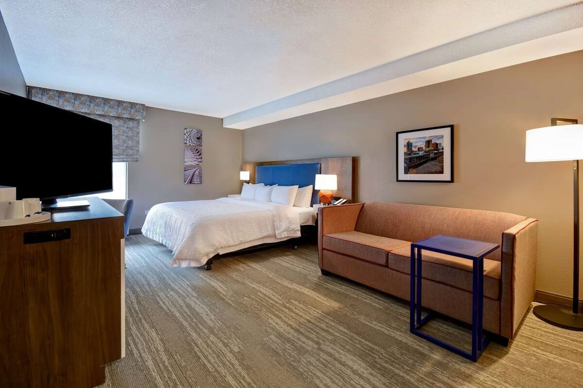 A hotel room with a bed, a sofa, a desk with a TV, and a lamp.