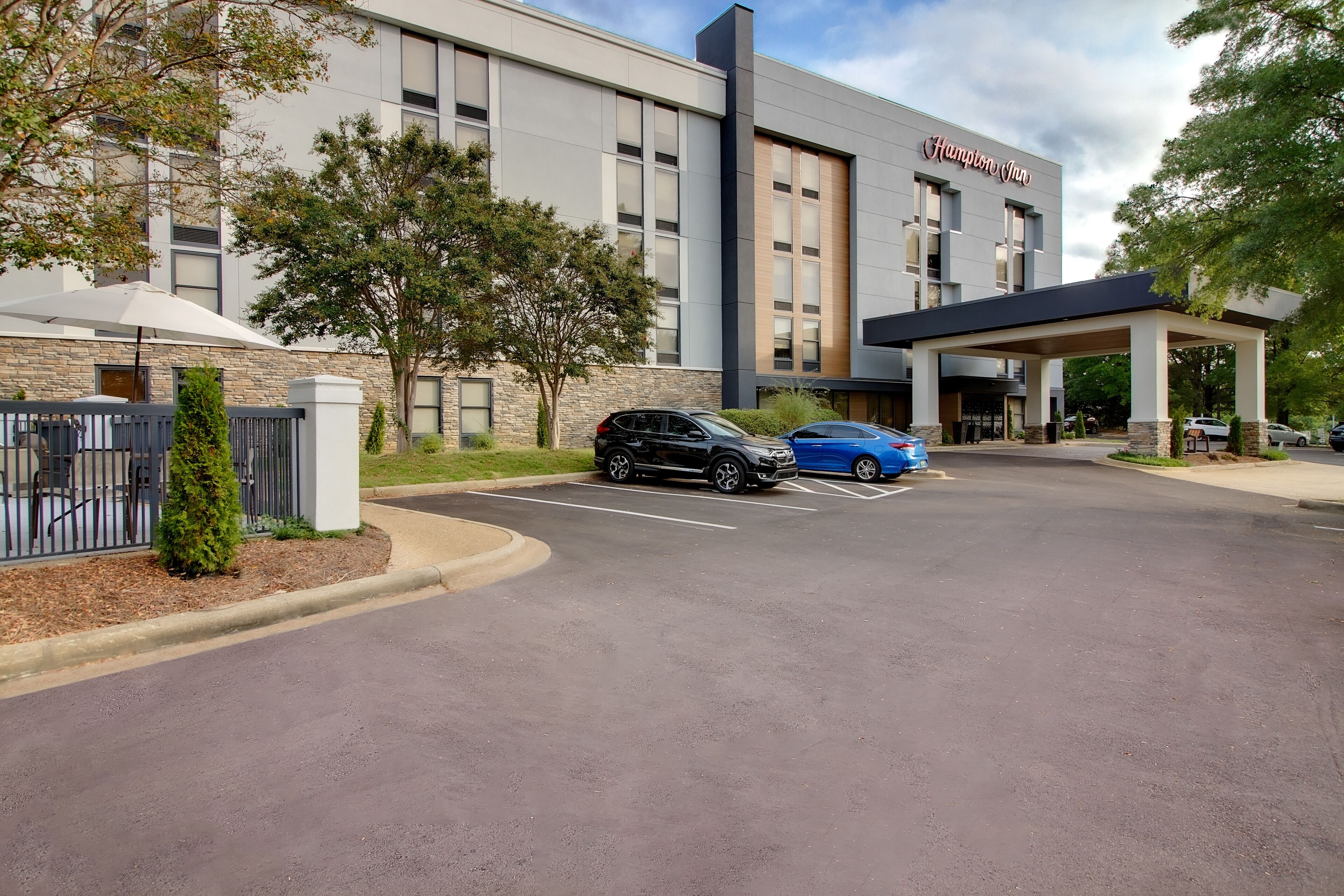 A modern hotel with a covered entrance, a stone facade, and a parking lot with several cars.