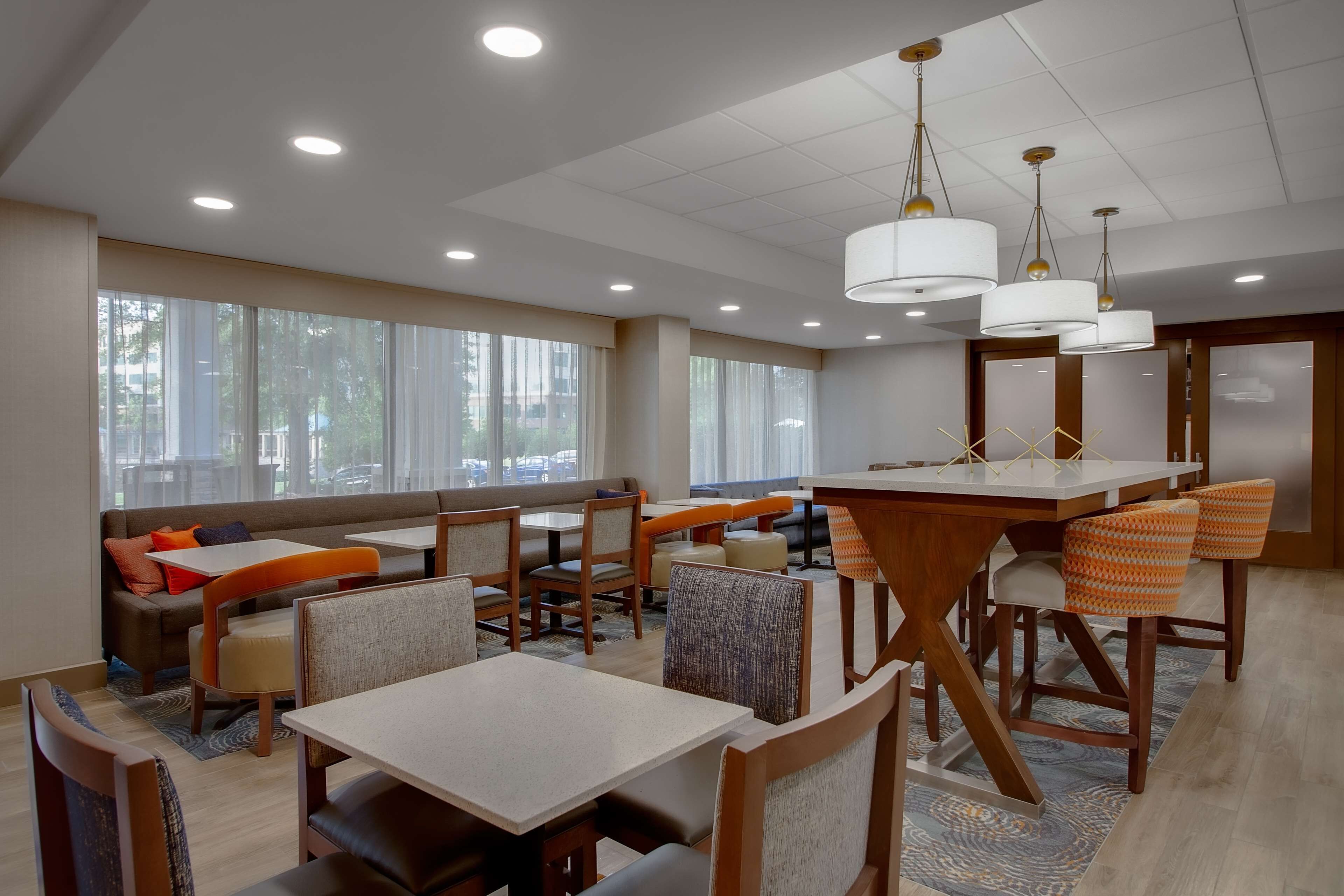 A modern hotel dining area with tables and chairs, large windows, and ceiling lights.