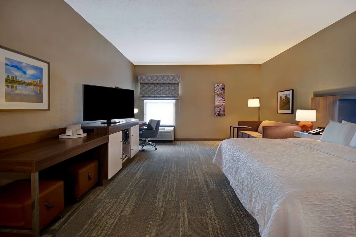 A hotel room with a bed, desk, television, and a window with a view of a cityscape.