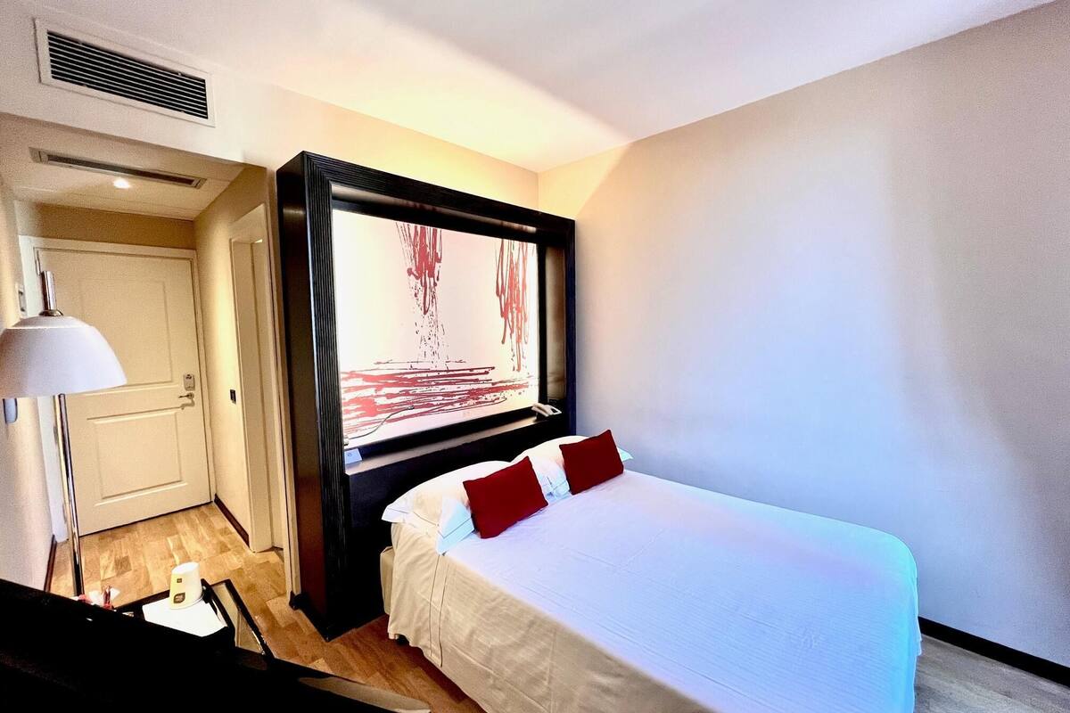 Une chambre d’hôtel avec un grand lit, un téléviseur à écran plat fixé au mur, un bureau avec une lampe et une porte donnant sur une autre pièce.
