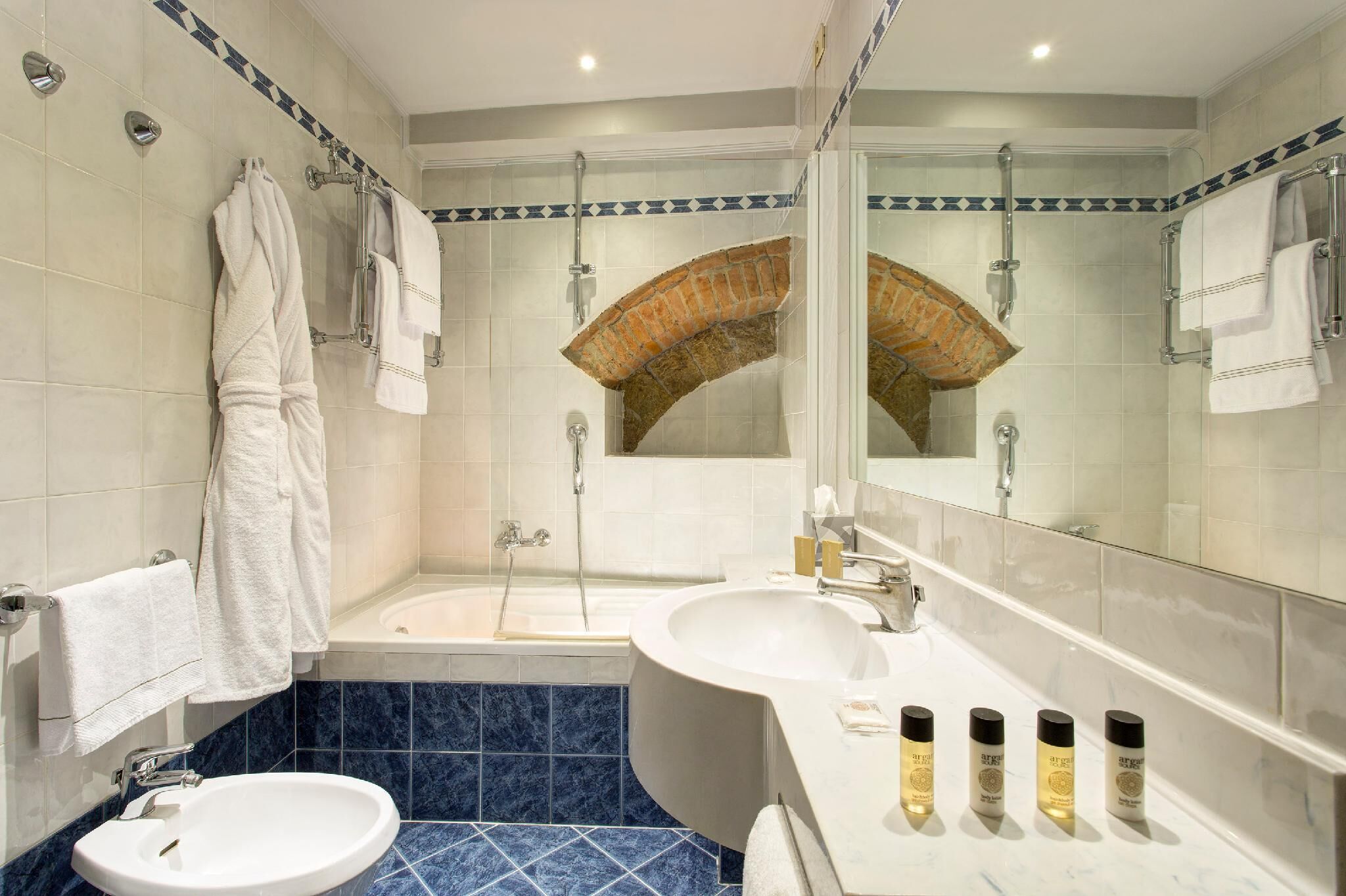 Un baño con bañera, lavamanos y un espejo grande. Hay toallas colgadas en un perchero y una ducha sobre la bañera.