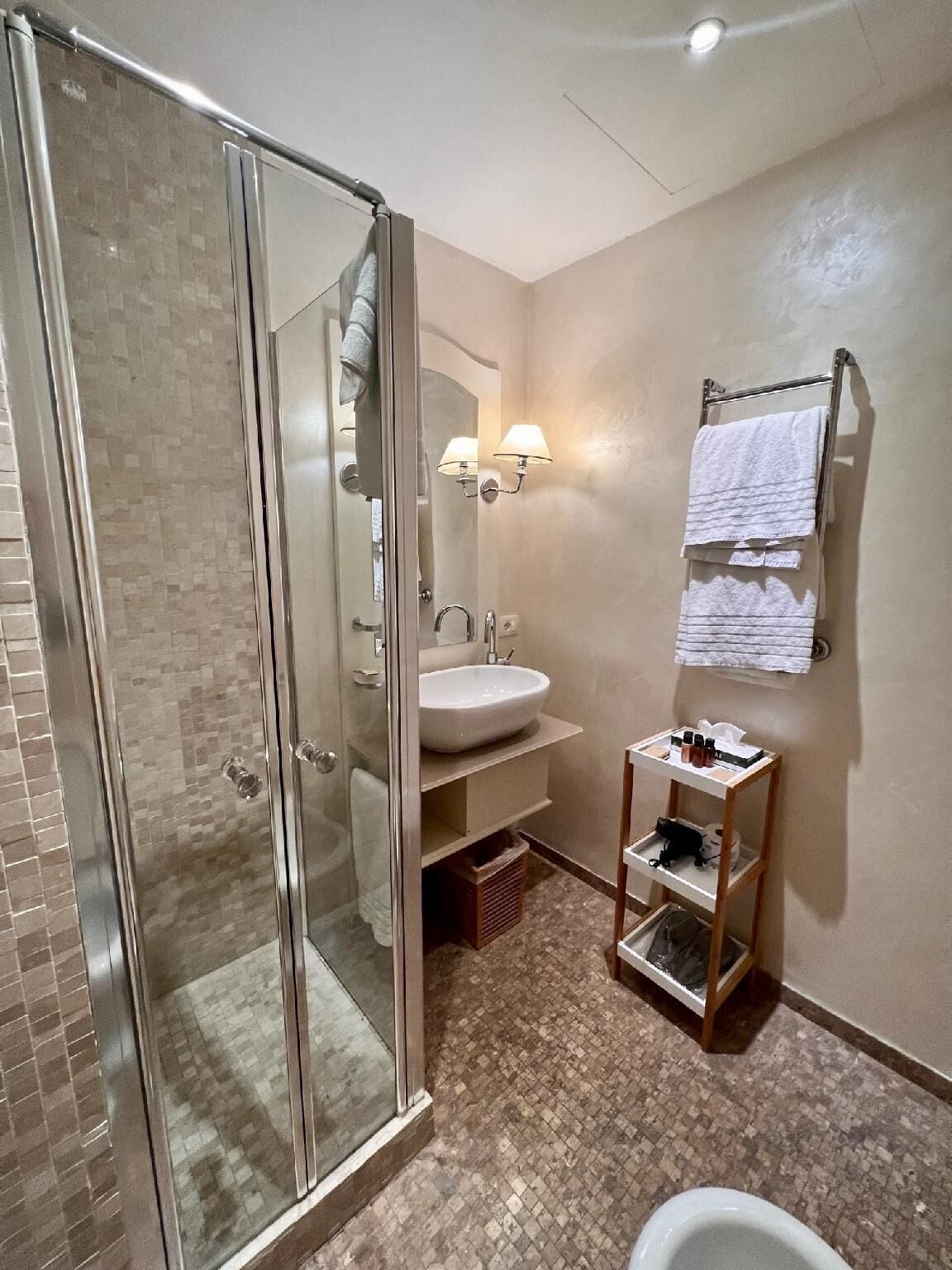 A bathroom with a glass shower enclosure, a sink, a mirror, and a small shelf with toiletries.