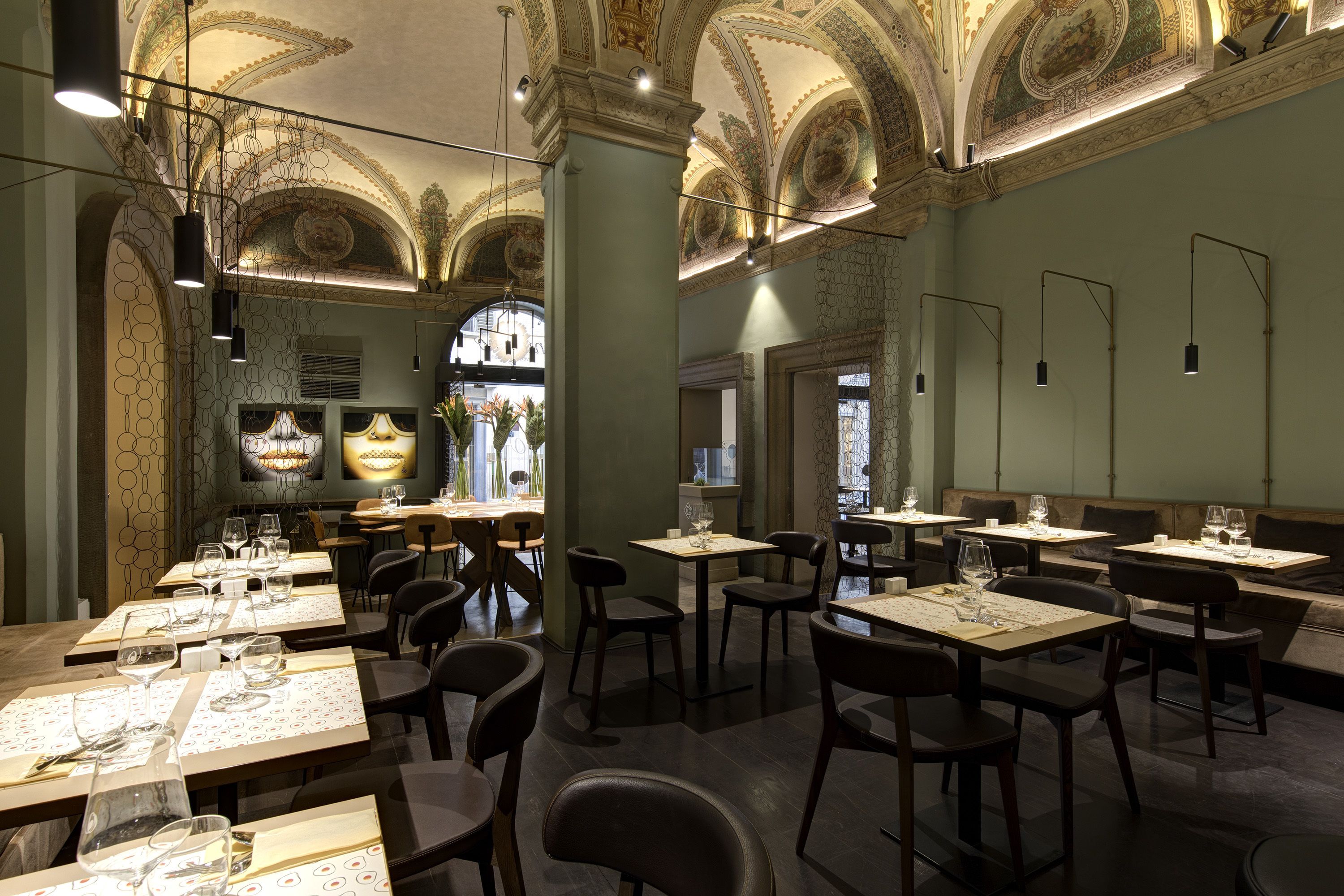 A restaurant with high ceilings, ornate ceiling decorations, and a warm, inviting atmosphere. Tables are set with wine glasses and menus, ready for guests.