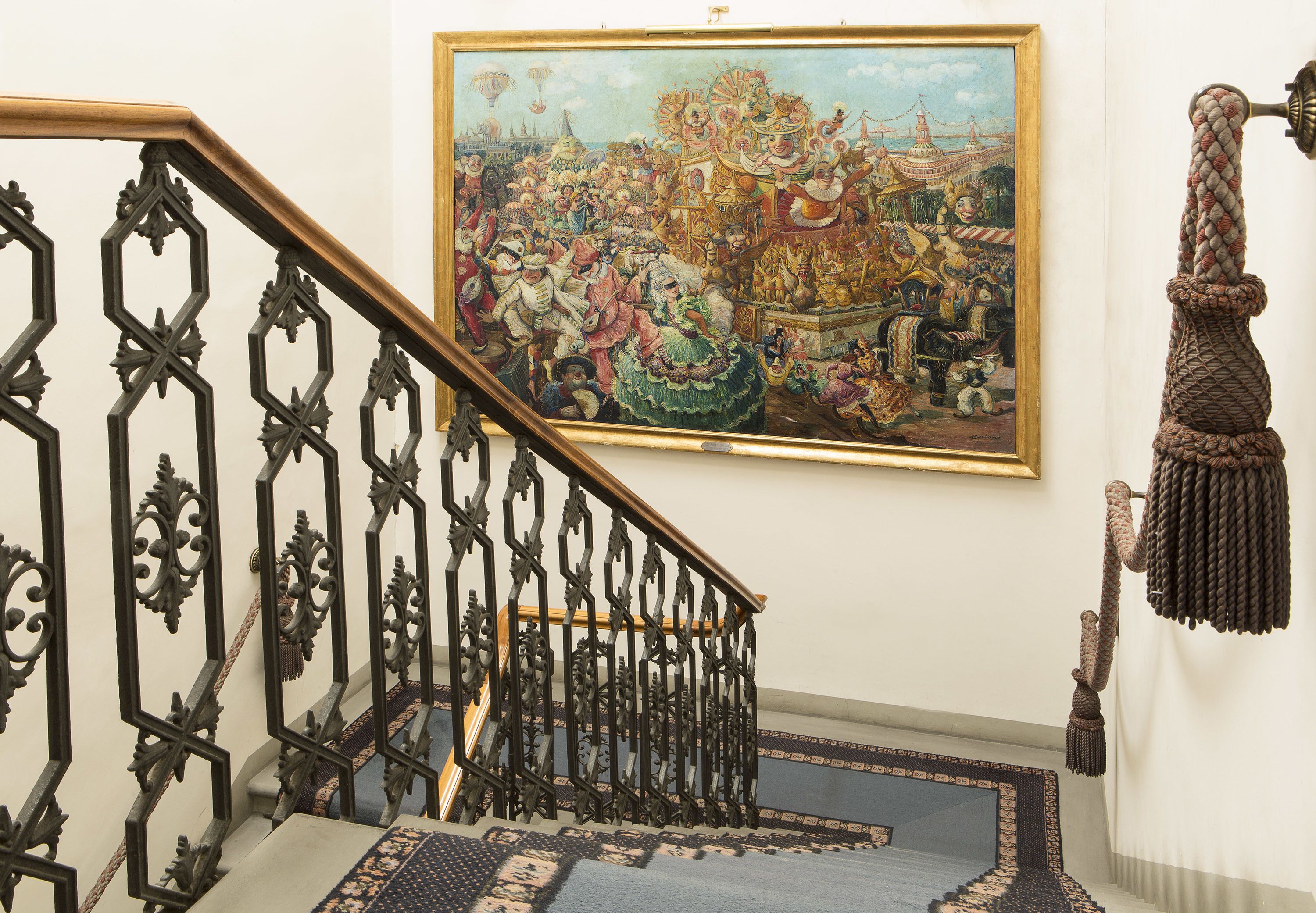 Eine Treppe mit aufwendiger schwarzer Metallgeländerung, einem gemusterten Teppich und einem großen, gerahmten Gemälde einer festlichen Szene an der Wand.