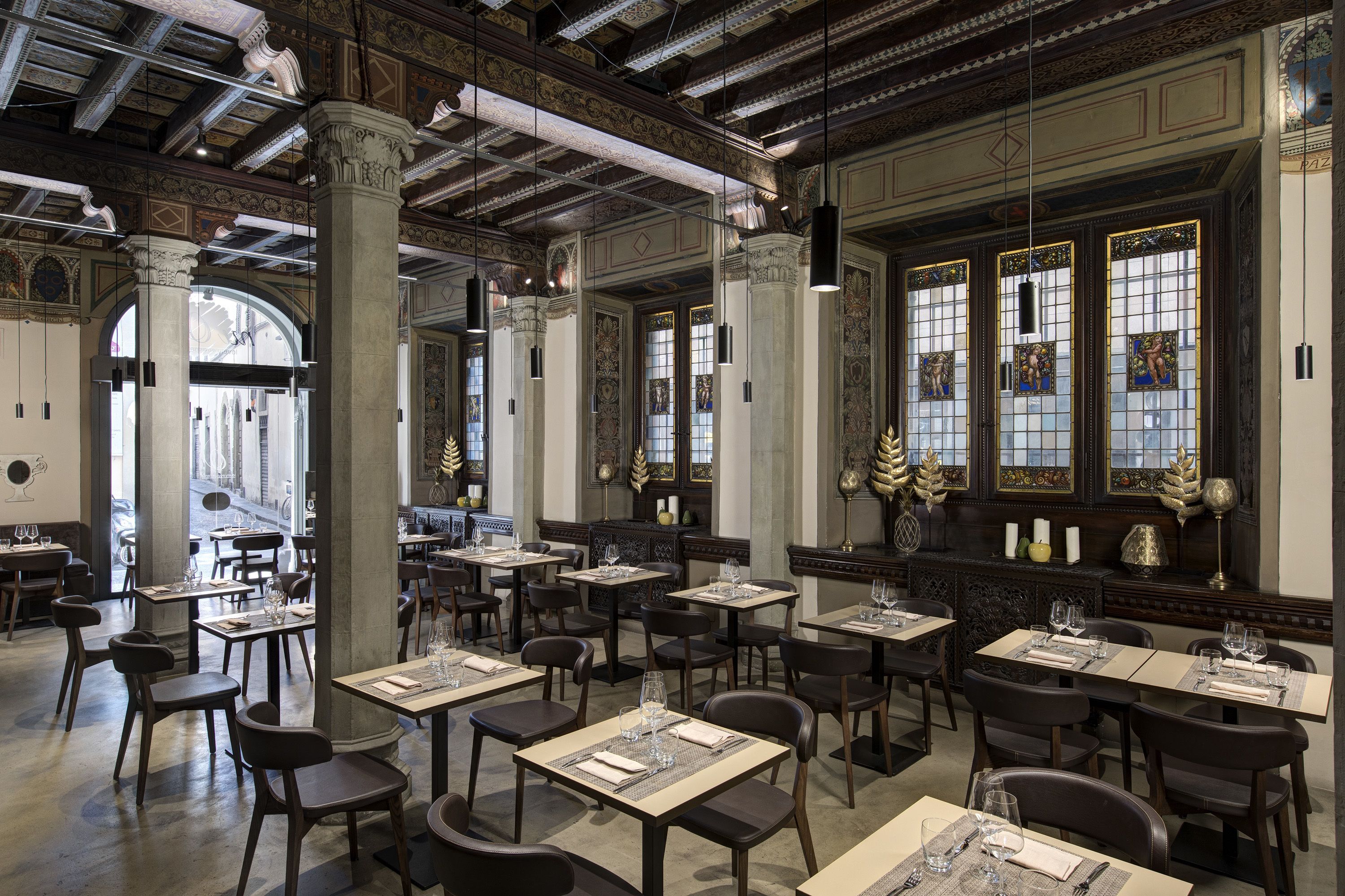 Un restaurant spacieux avec de hauts plafonds, des colonnes richement décorées et de grandes fenêtres ornées de vitraux. Les tables sont disposées avec soin et accompagnées de chaises, et l’intérieur présente un style architectural classique.