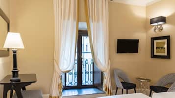 Ein Hotelzimmer mit einem Bett, einem Schreibtisch, zwei Stühlen, einem kleinen Tisch, einer Lampe, einem Fernseher und einem Balkon mit Vorhängen.