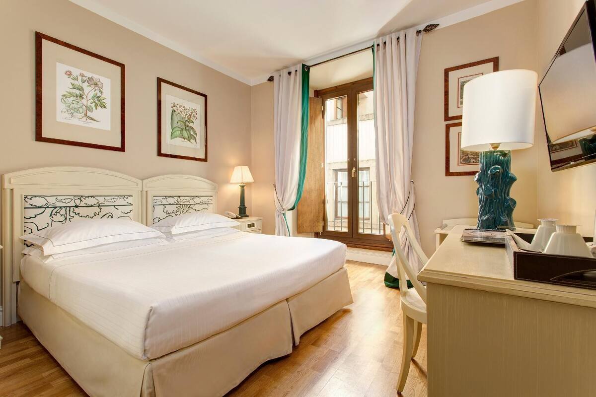 Une chambre d’hôtel avec un grand lit, un bureau avec une lampe et des reproductions encadrées de motifs botaniques accrochées au mur.