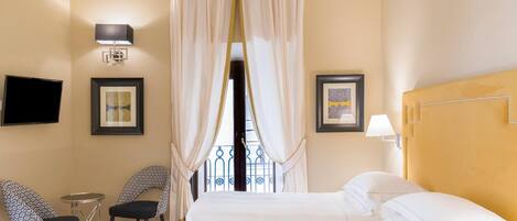 A hotel room with a bed, two chairs, a small table, a TV, and a window with curtains.