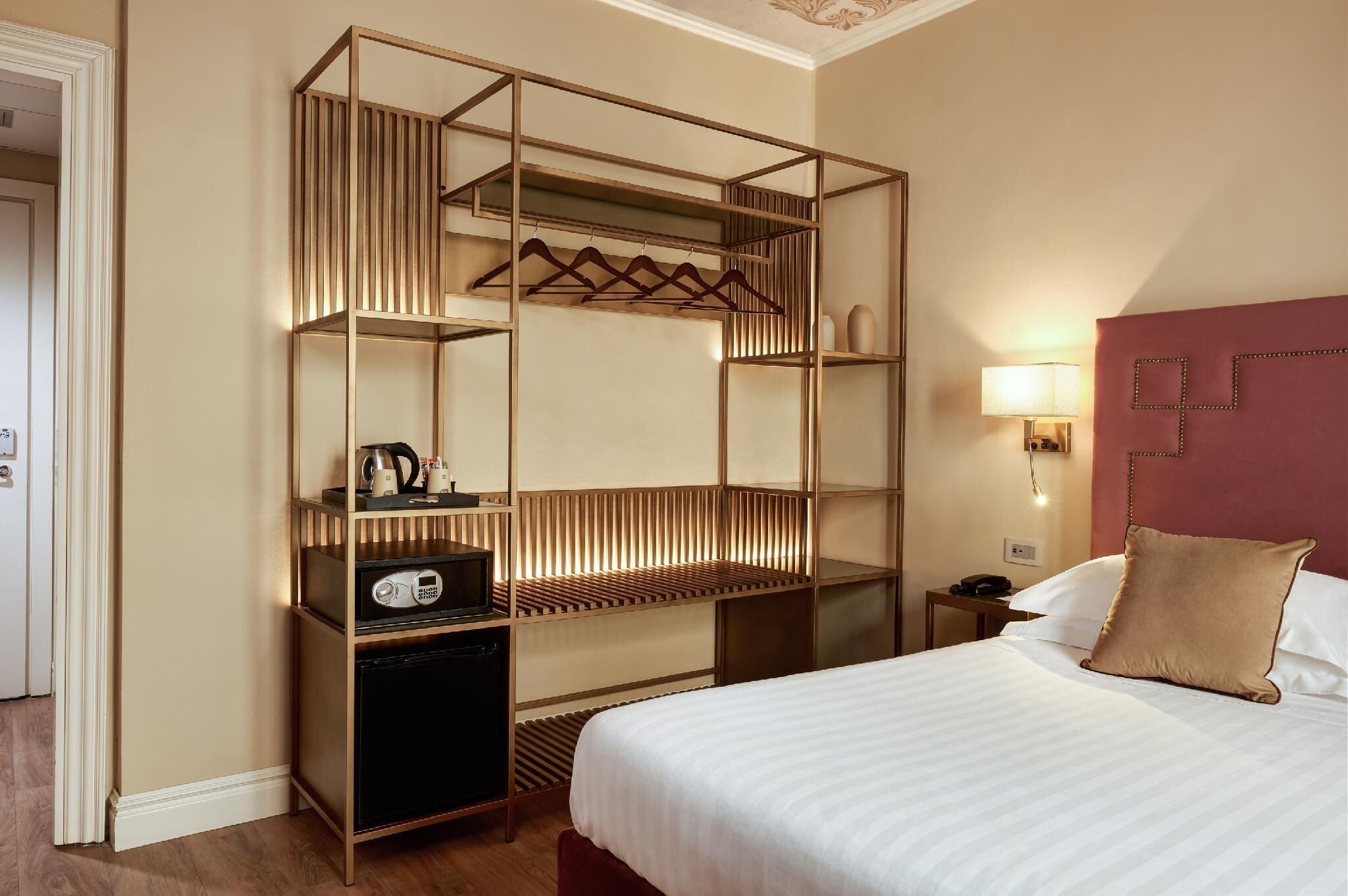 A hotel room with a bed, a bedside table with a lamp, a wardrobe with hanging clothes, and a small desk with a radio.