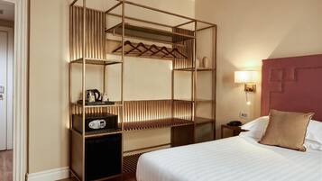 A hotel room with a bed, a bedside table with a lamp, a wardrobe with hanging clothes, and a small desk with a radio.