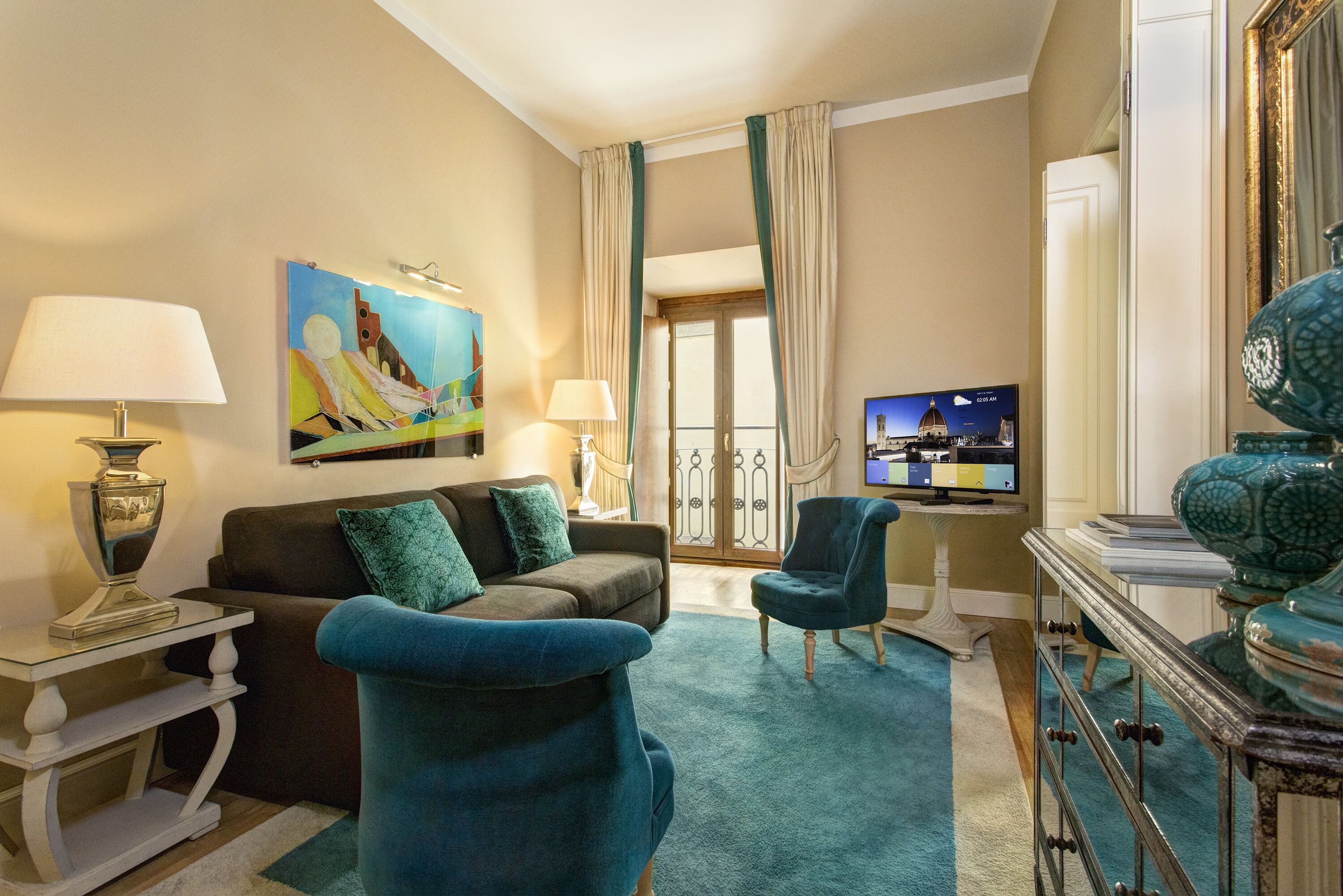 A hotel room with a sofa, two armchairs, a TV, a painting, and a view of the cityscape through the window.
