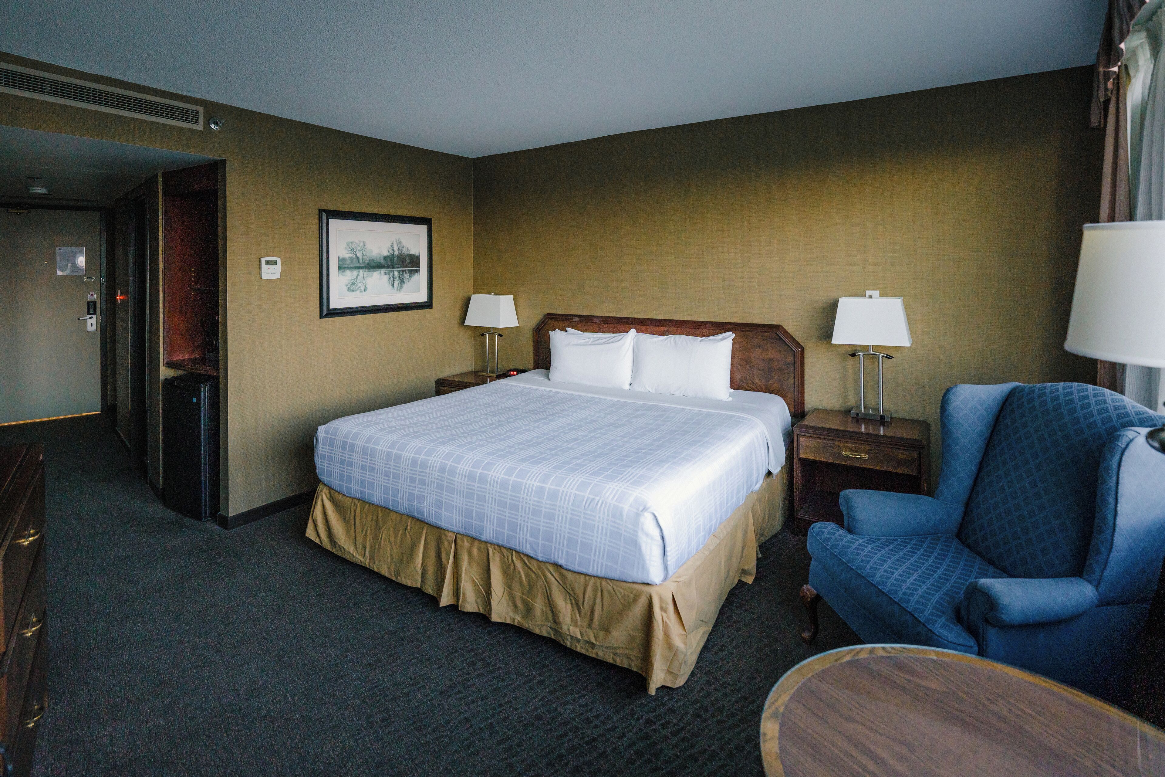 A hotel room with a large bed, two bedside tables with lamps, a blue armchair, and a small wooden table.