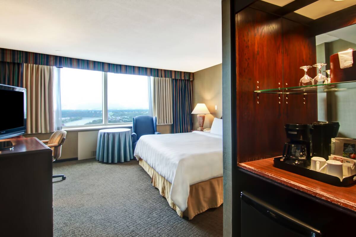A hotel room with a bed, a desk with a TV, a chair, a small table, a window with curtains, and a coffee station.