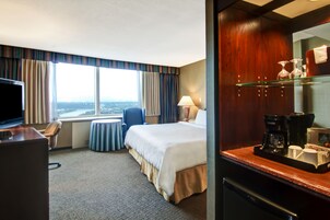 A hotel room with a bed, a desk with a TV, a chair, a small table, a window with curtains, and a coffee station.