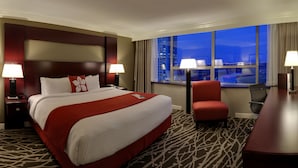 A hotel room with a large bed, two bedside tables with lamps, a red chair, a desk, and a large window with a city view. - Chateau Lacombe Hotel (Edmonton)