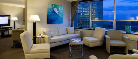Ein Hotelzimmer mit einer Couch, Sesseln, einem Couchtisch, einem Fernseher und einem großen Fenster mit Blick auf die Stadt.