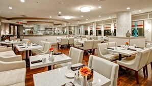 Un restaurante moderno con mesas y sillas blancas, una zona de bar central y una columna grande.