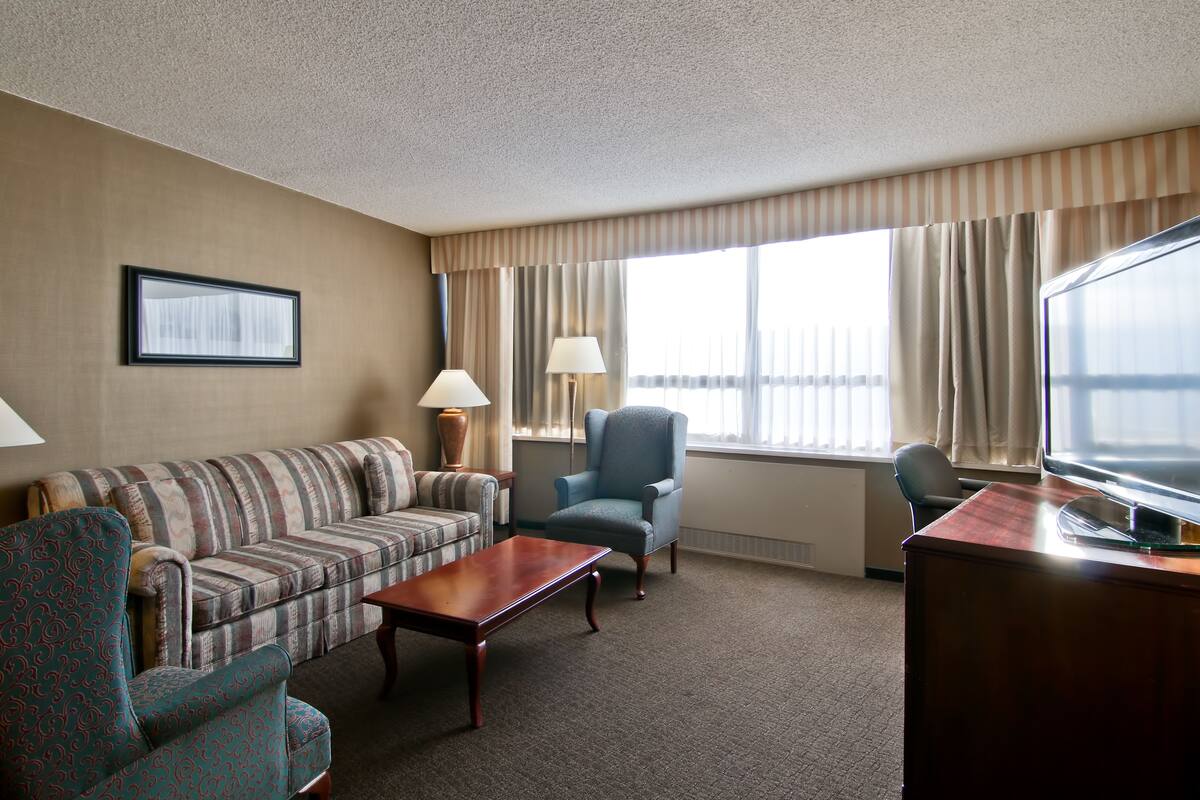 A hotel room with a striped sofa, a blue armchair, a wooden coffee table, a TV, and a large window with curtains.