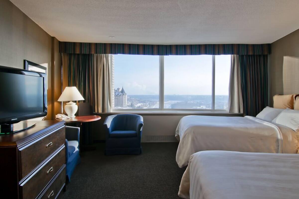 A hotel room with a large window, a TV, a desk with a lamp, and two beds.