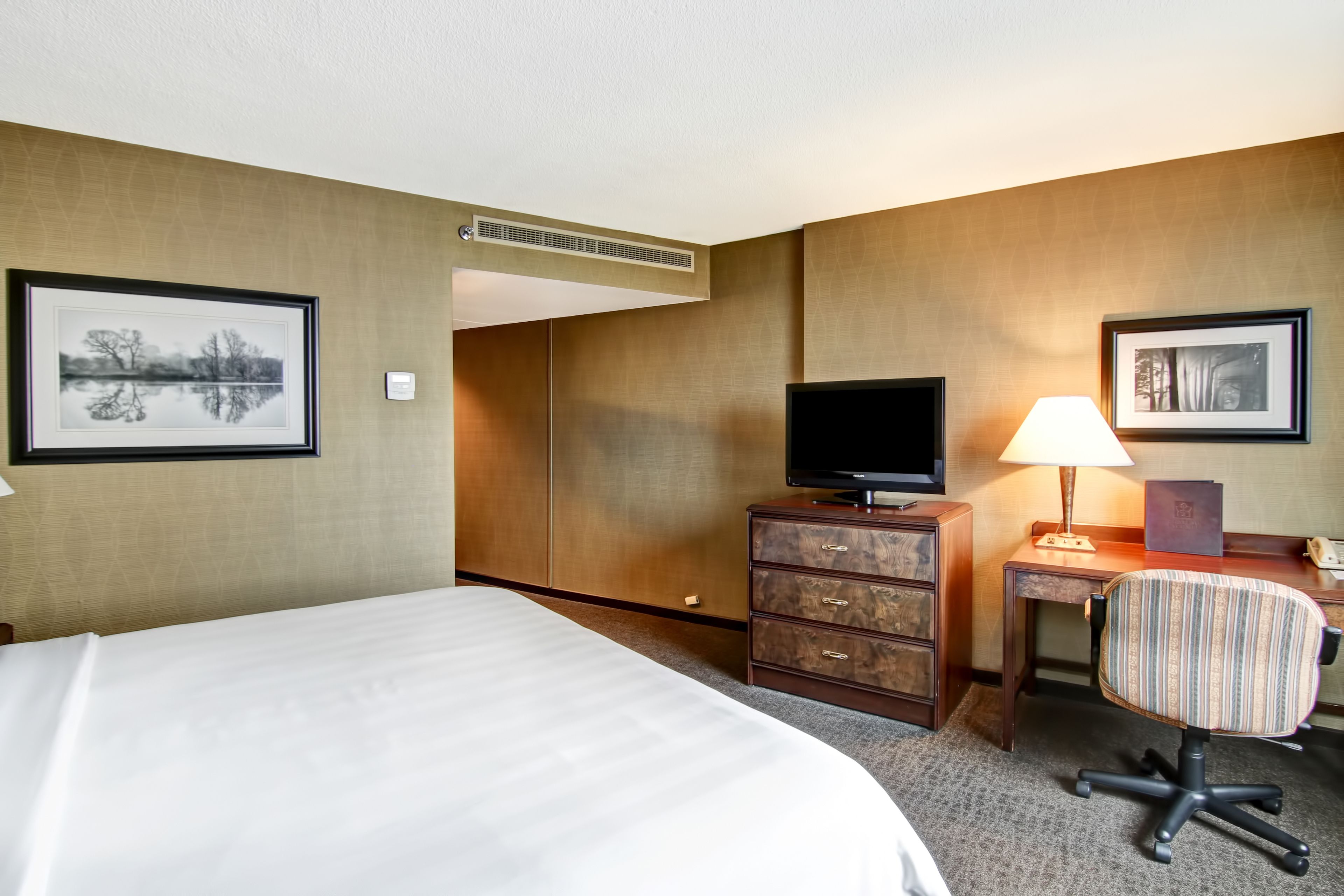 A hotel room with a bed, a desk with a chair, a television, a lamp, and two framed pictures on the walls.