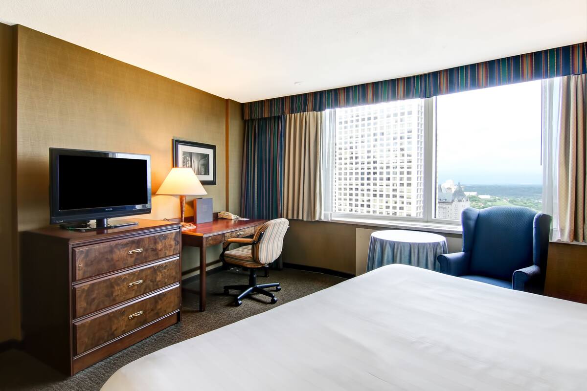 A hotel room with a bed, desk, chair, television, lamp, and a window with a city view.
