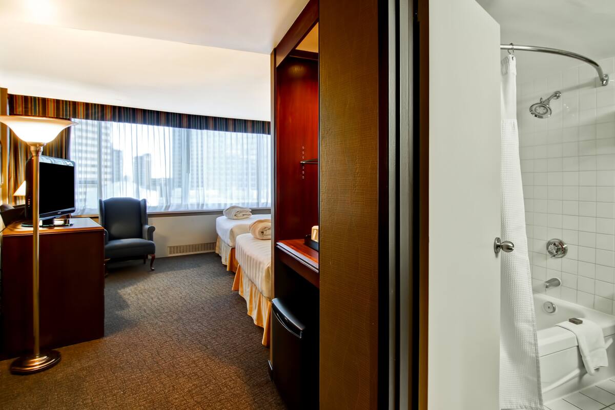 A hotel room with a desk, chair, two beds, a shower, and a view of buildings through the window.
